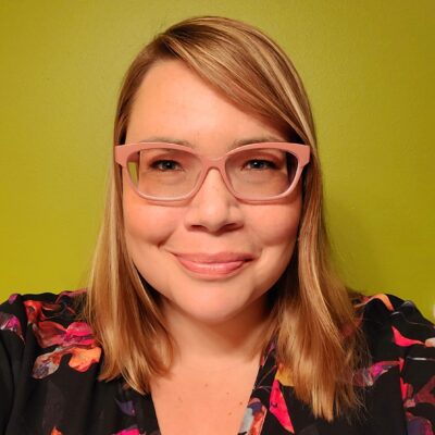 Photo of Anna Hildebrandt, a woman with light, shoulder-length hair wearing pink eyeglasses